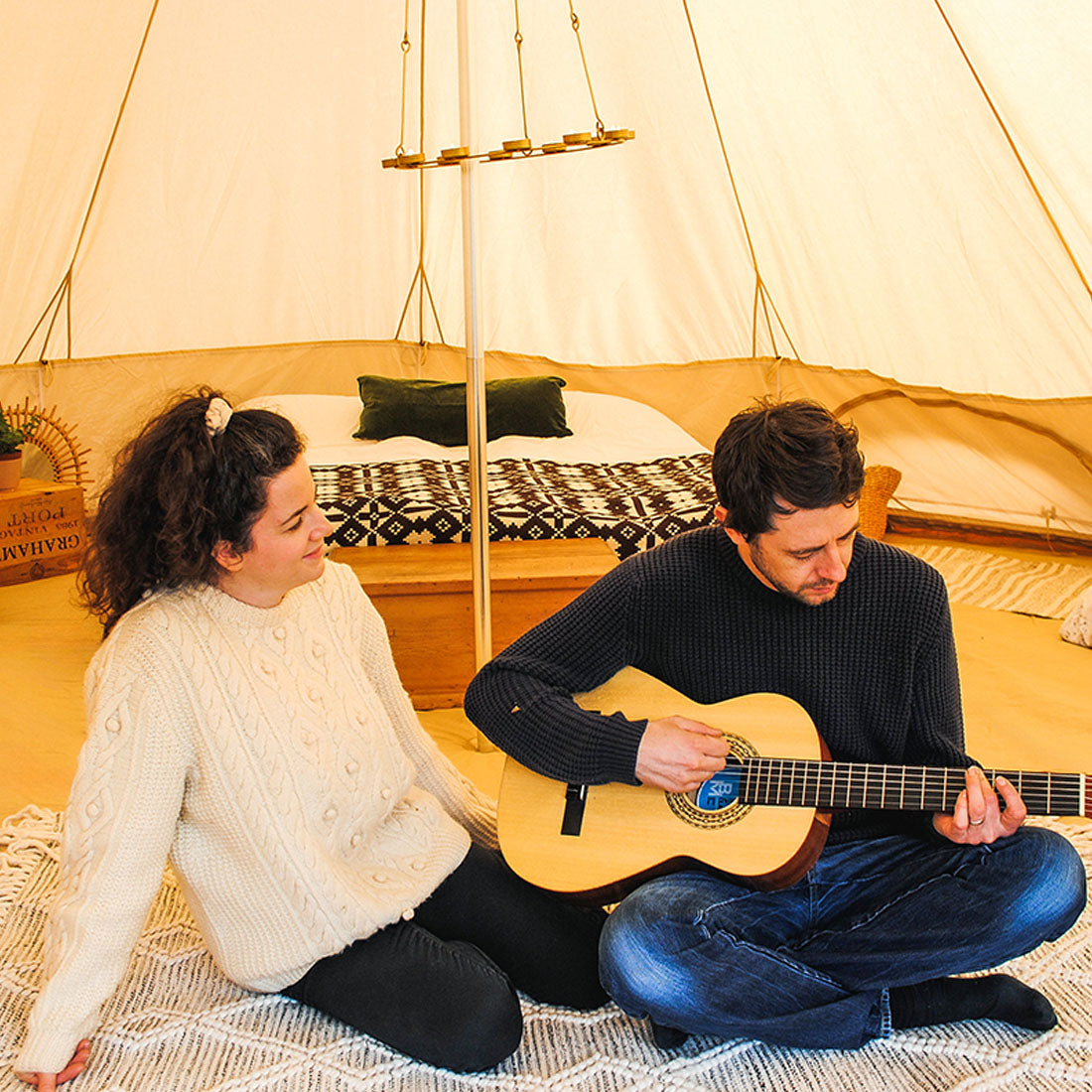 a couple playing the guitar sitting on a rug inside a boutique camping star bell tent, a double bed and chandelier are in the background