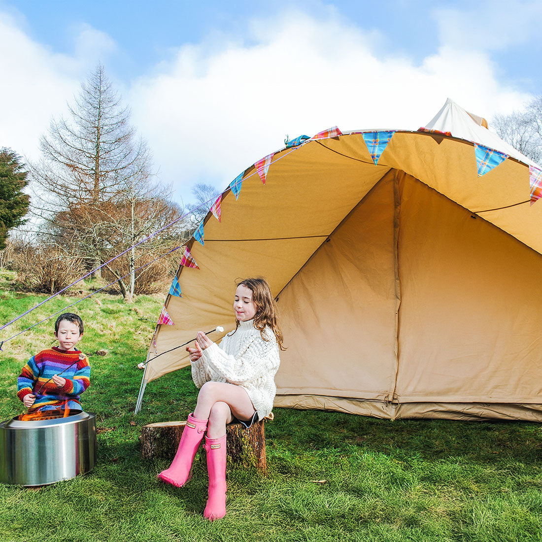 a girl and a boy sitting on logs toasting and eating marshmallows outside a sandstone star bell tent in a green field