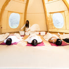 A woman sits inside a boutique camping nova air dome glamping tent, holding white bowls while three people lie on pink yoga mats on the floor, wrapped in white blankets.