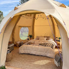 A boutique camping nova air dome glamping tent with a fully furnished bedroom inside, featuring a bed with beige bedding, a woven rug, string lights, and a window looking out onto a green landscape under a blue sky.