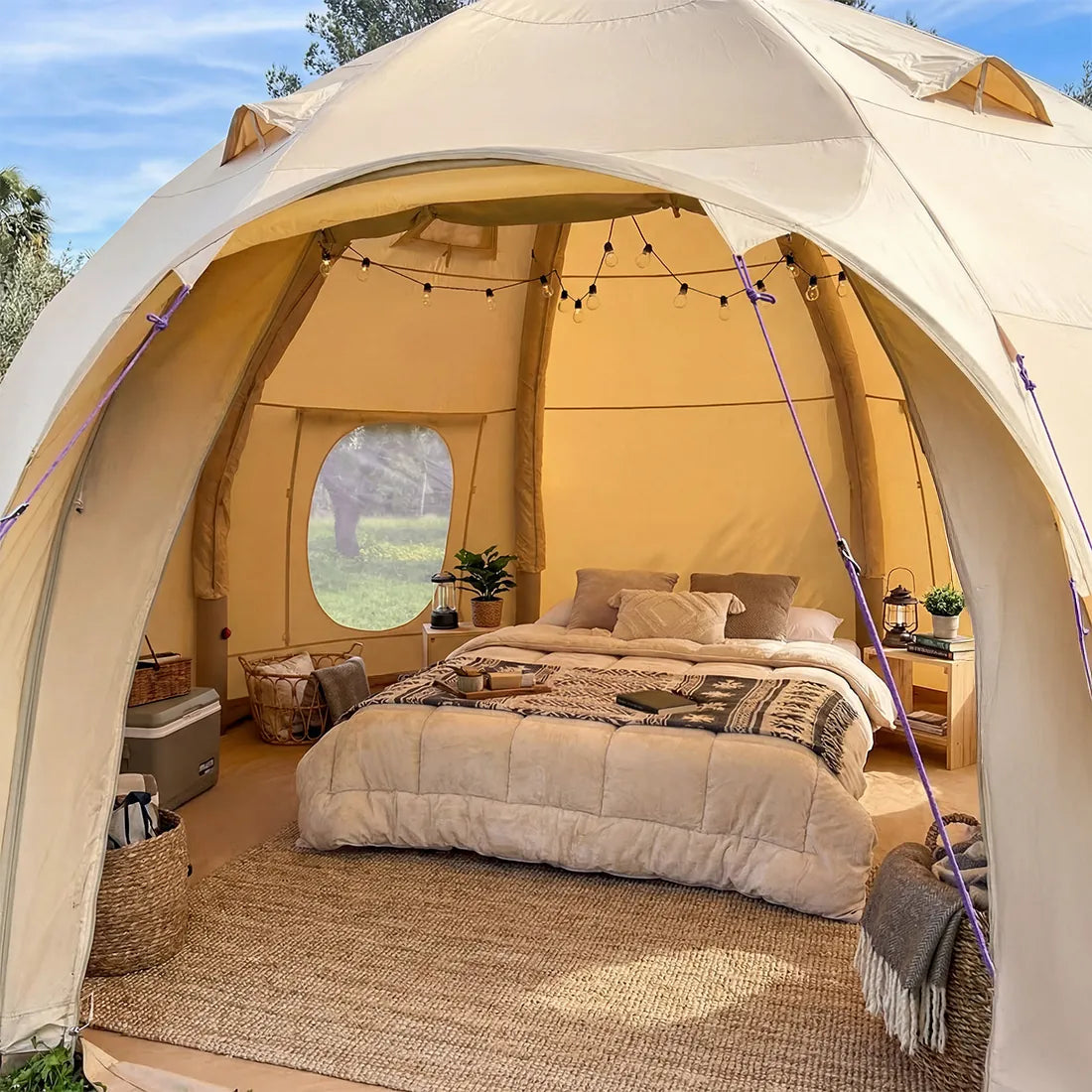 A boutique camping nova air dome glamping tent with a fully furnished bedroom inside, featuring a bed with beige bedding, a woven rug, string lights, and a window looking out onto a green landscape under a blue sky.