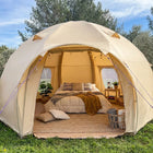 A boutique camping nova air dome glamping tent pitched outdoors on grass with trees and blue sky in the background. The tent is open, revealing a styled bed inside with patterned pillows, a brown throw blanket, a small side table, and a woven basket holding folded blankets.