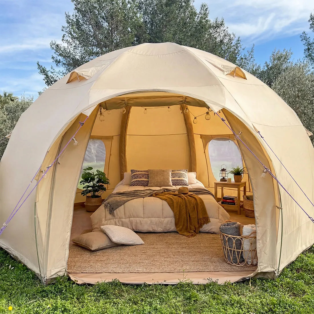 A boutique camping nova air dome glamping tent pitched outdoors on grass with trees and blue sky in the background. The tent is open, revealing a styled bed inside with patterned pillows, a brown throw blanket, a small side table, and a woven basket holding folded blankets.