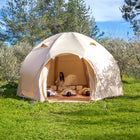 A boutique camping nova air dome glamping tent in beige is pitched on a grassy lawn outdoors, with trees and blue sky in the background. A woman is visible sitting inside the open tent, and bedding or cushions are arranged on the floor.