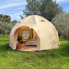 A boutique camping nova air dome glamping tent pitched on a grassy lawn outdoors, with trees and sky in the background. The tent is open, showing a furnished interior with a bed, pillows, and string lights.