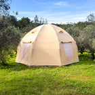 A boutique camping nova air dome glamping in light beige is pitched on a grassy field with olive trees and a blue sky in the background.