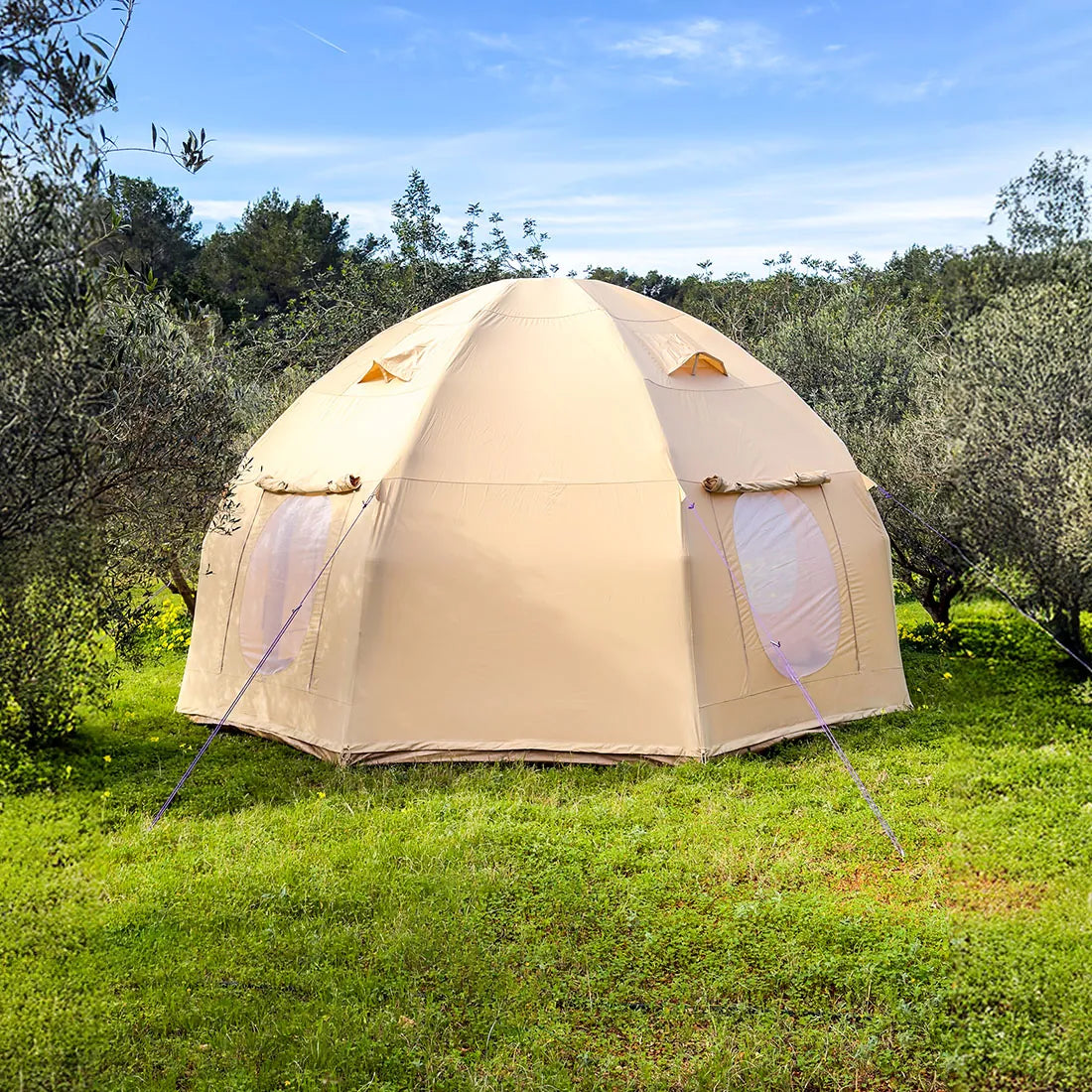 A boutique camping nova air dome glamping in light beige is pitched on a grassy field with olive trees and a blue sky in the background.