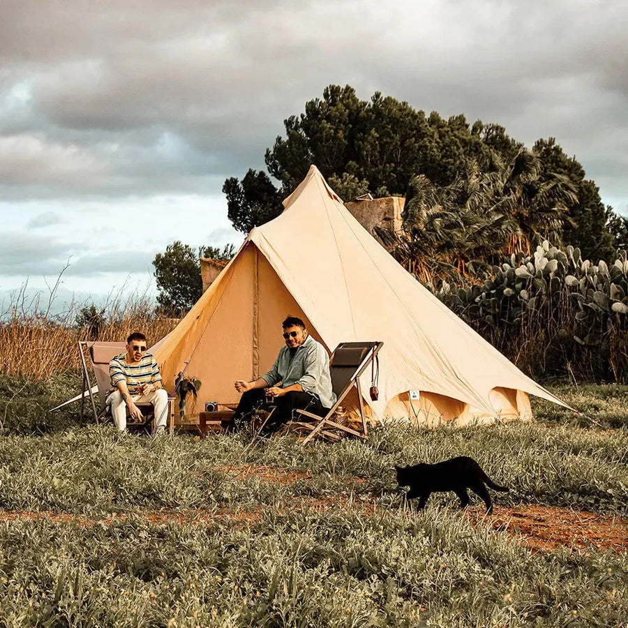 Two people sitting in front of the Bell Tent Plus in a natural setting with a black cat walking nearby.