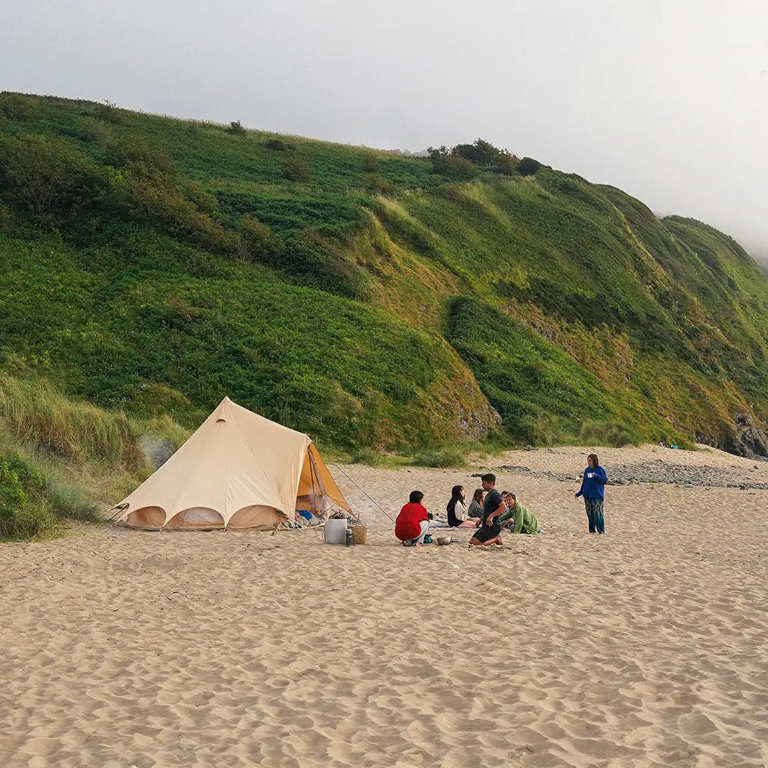 Group of people camping on a sandy beach with a bell tent plus glamping tent and a scenic hillside in the background.