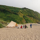 Group of people camping on a sandy beach with a bell tent plus glamping tent and a scenic hillside in the background.