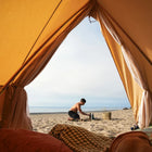 Person setting up camp on a sandy beach from inside an bell tent plus glamping tent by boutique camping 