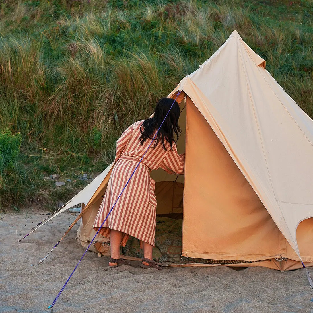 Person in a striped dress standing next to a boutique camping bell tent plus glamping tent on a sandy beach.