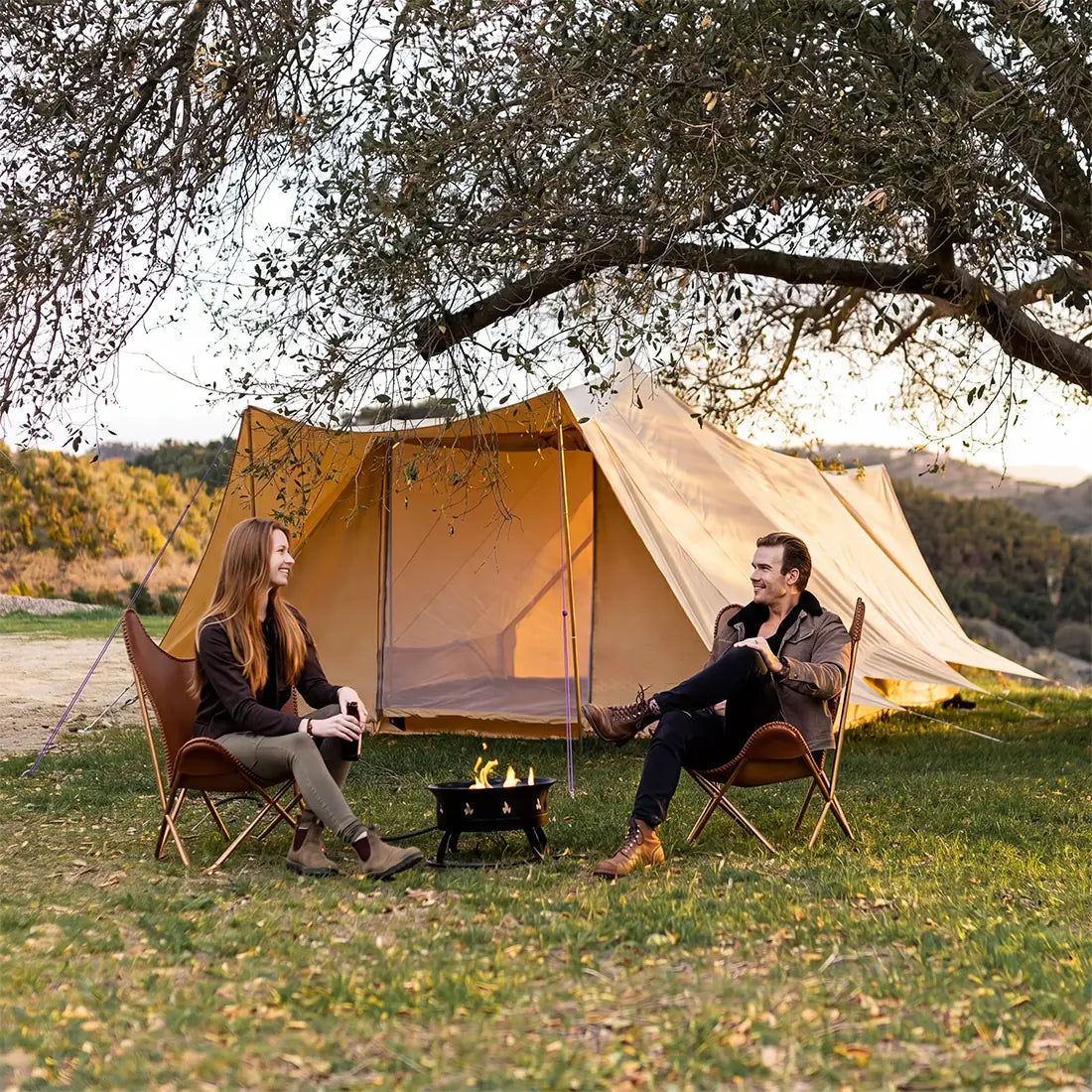 a couple sitting in front of a boutique camping tucana double door bell tent in a natural setting with a small fire pit 