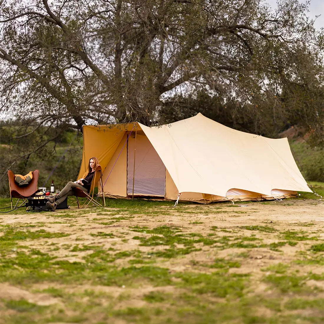 Person sitting near a boutique camping tucana double door tent in a natural setting with trees and grass.