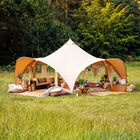 A boutique camping phoenix tent set up in a grassy field with trees in the background, surrounded by lounge chairs, cushions, tables, and string lights.