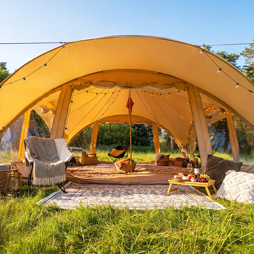 boutique camping phoenix tent set up outdoors in a grassy field under a blue sky, furnished with a chair, blanket, pillows, and a small table.