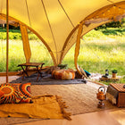 A cozy boutique camping phoenix glamping tent interior with a warm yellow canopy, woven rugs, colorful patterned pillows, a small folding table, string lights, and an outdoor grassy setting visible through the tent openings.