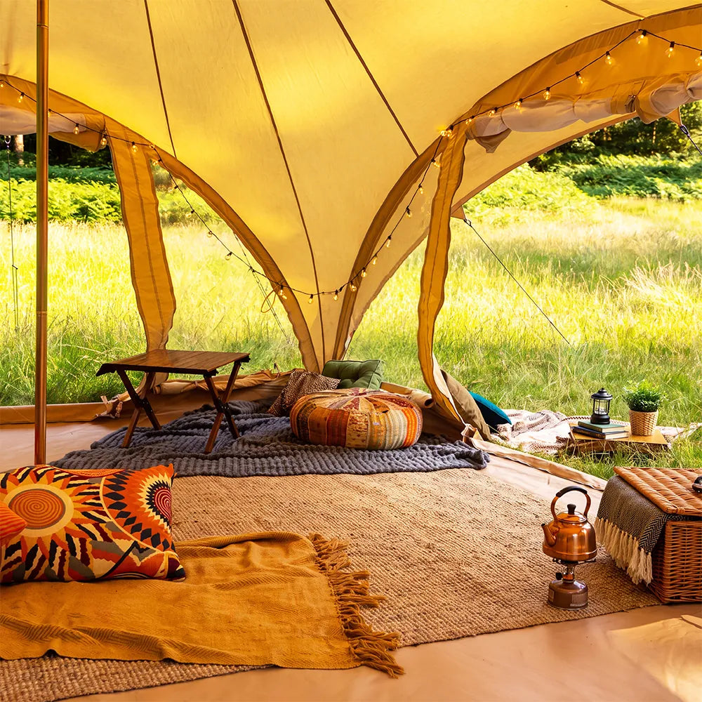 A cozy boutique camping phoenix glamping tent interior with a warm yellow canopy, woven rugs, colorful patterned pillows, a small folding table, string lights, and an outdoor grassy setting visible through the tent openings.