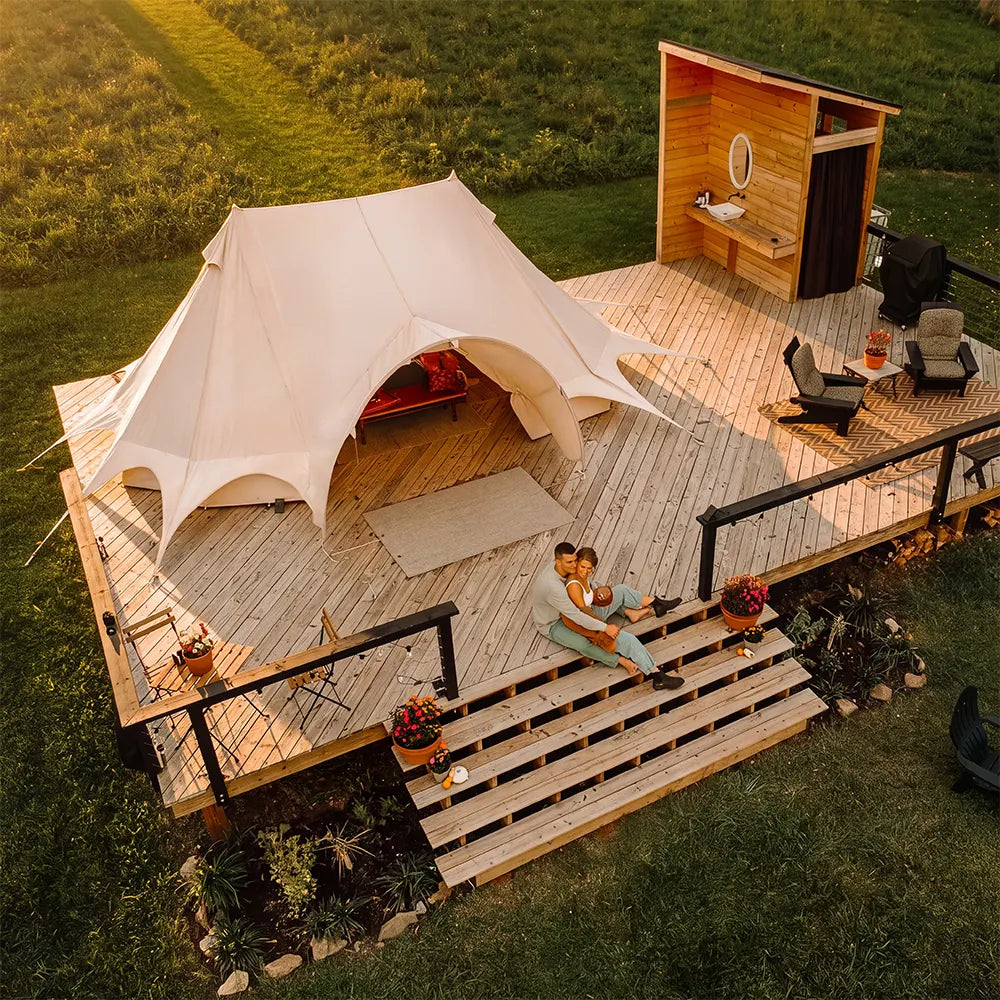 An aerial view of a luxury glamping setup featuring a boutique camping star emperor bell tent on a wooden deck, a small wooden cabin with a bathroom, and a couple sitting on the deck steps in the warm glow of sunset.