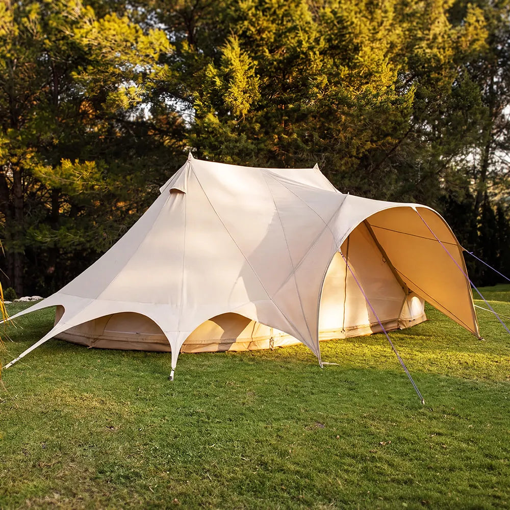 A boutique camping star emperor bell tent pitched on a grassy lawn with trees in the background, showing one end open to the air.