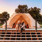 A boutique camping star emperor bell tent with a curved roof is set on a wooden platform outdoors at sunset. A woman wearing a yellow beanie and plaid shirt sits inside the tent next to a fluffy light-colored dog.