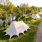 A boutique camping star emperor bell tent with dramatic pointed starburst corners is pitched on a grassy lakeside campsite, with other similar tents, trees, and a gravel path in the background.