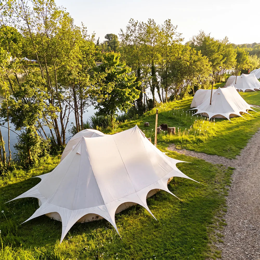 A boutique camping star emperor bell tent with dramatic pointed starburst corners is pitched on a grassy lakeside campsite, with other similar tents, trees, and a gravel path in the background.
